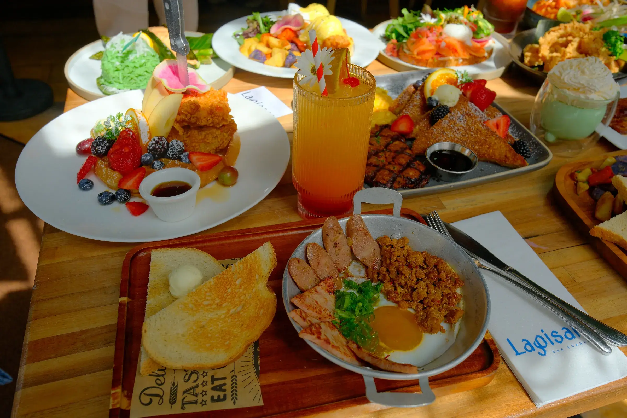 A wooden table of brunch plates with a rice pan, fried chicken and a sunrise iced drink